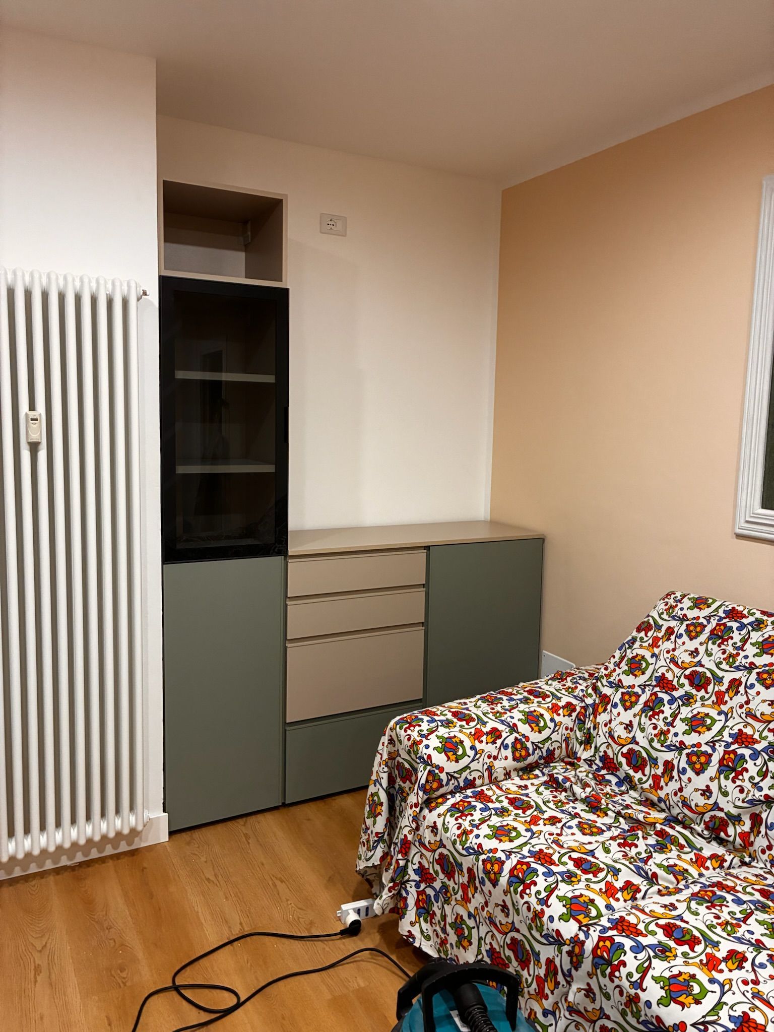 Camera da letto piccola con un letto, un armadio, un radiatore e un cavo di alimentazione nero sul pavimento.