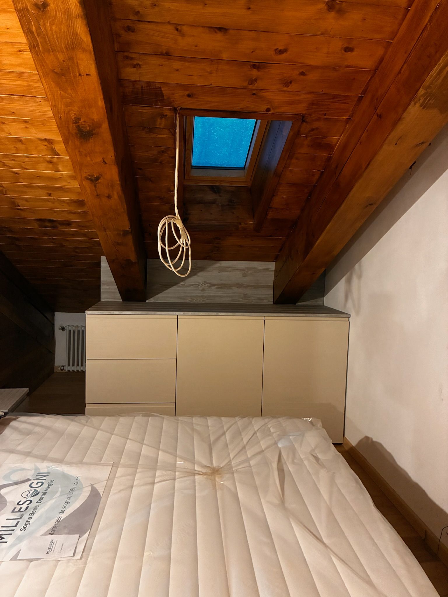 Camera da letto con soffitto spiovente in legno, lucernario, armadi beige e letto bianco in primo piano