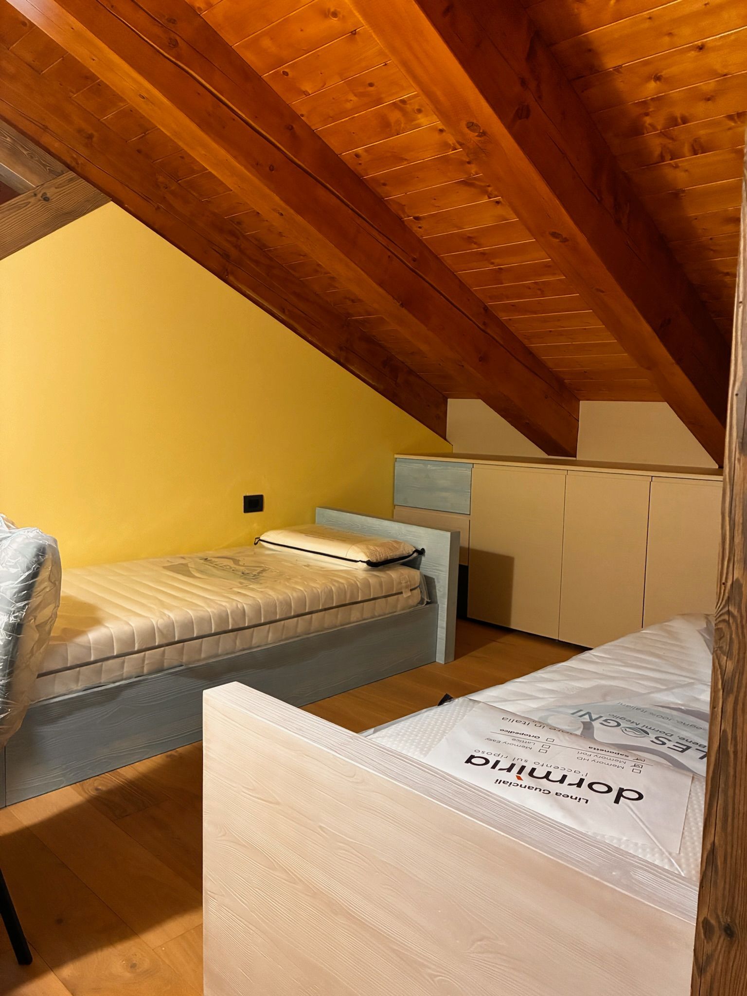Camera da letto in mansarda con soffitto spiovente in legno e due letti singoli, uno bianco e uno grigio, illuminata da una luce calda
