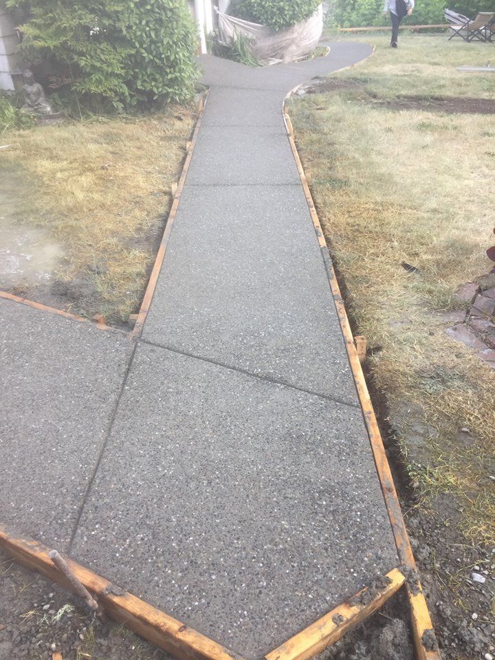 Narrow concrete garden path with dark edging, leading between grassy areas toward plants and steps