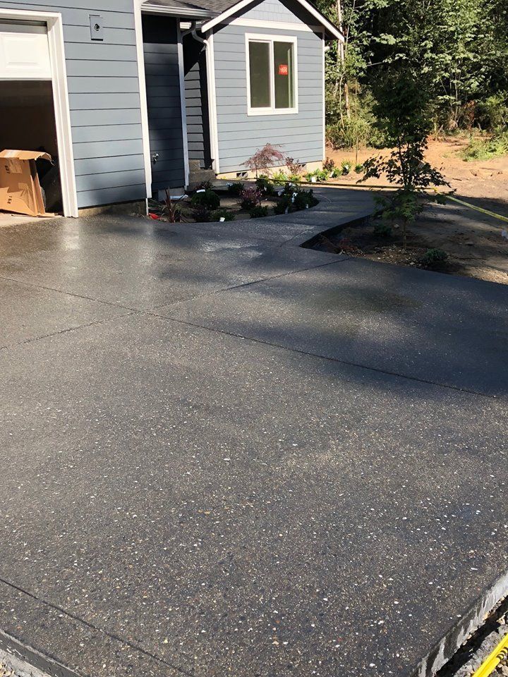 Freshly paved driveway beside a gray house and garage, with a small landscaped entrance and wooded yard.