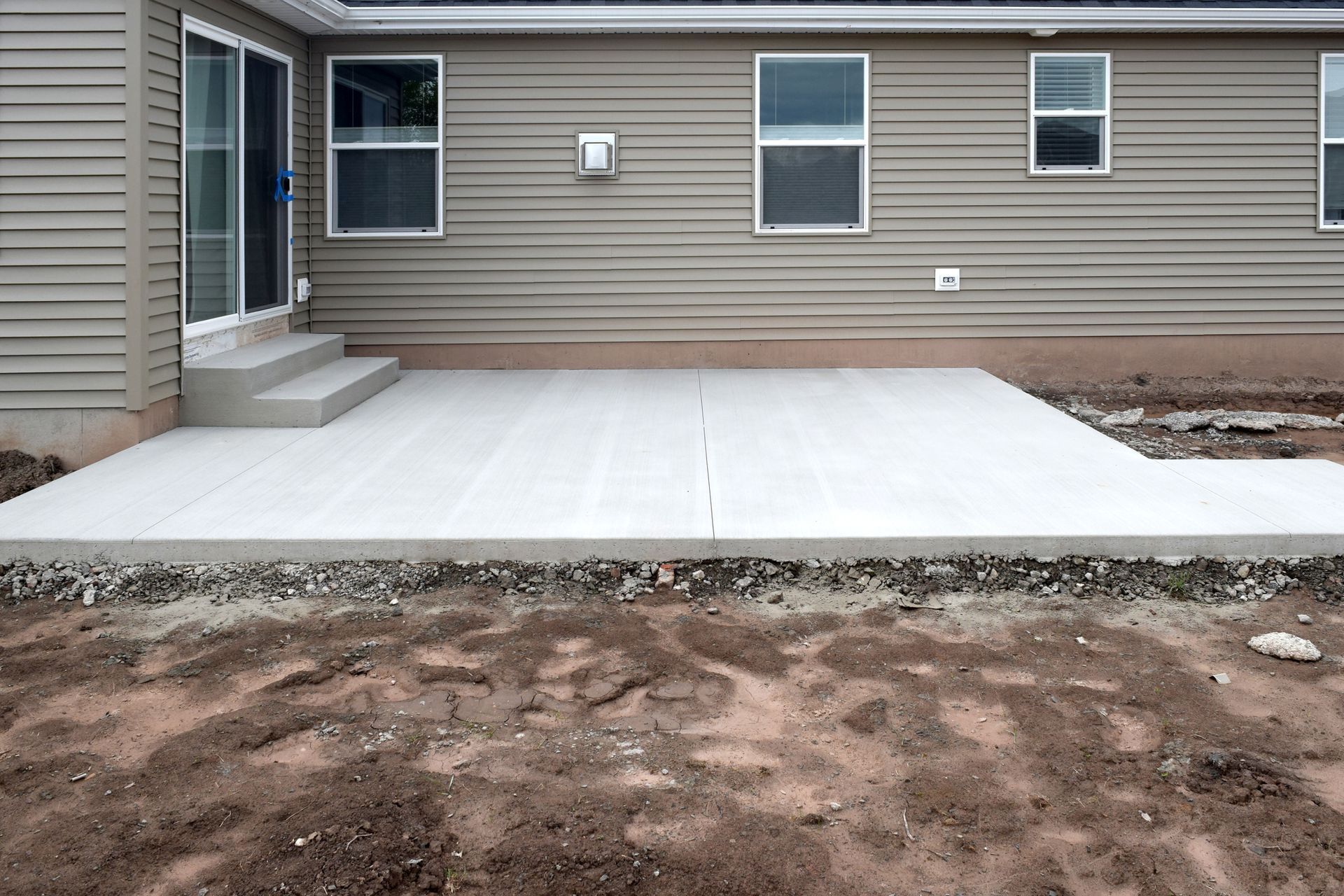 Fresh concrete patio beside a beige house, with steps and a dirt yard in front