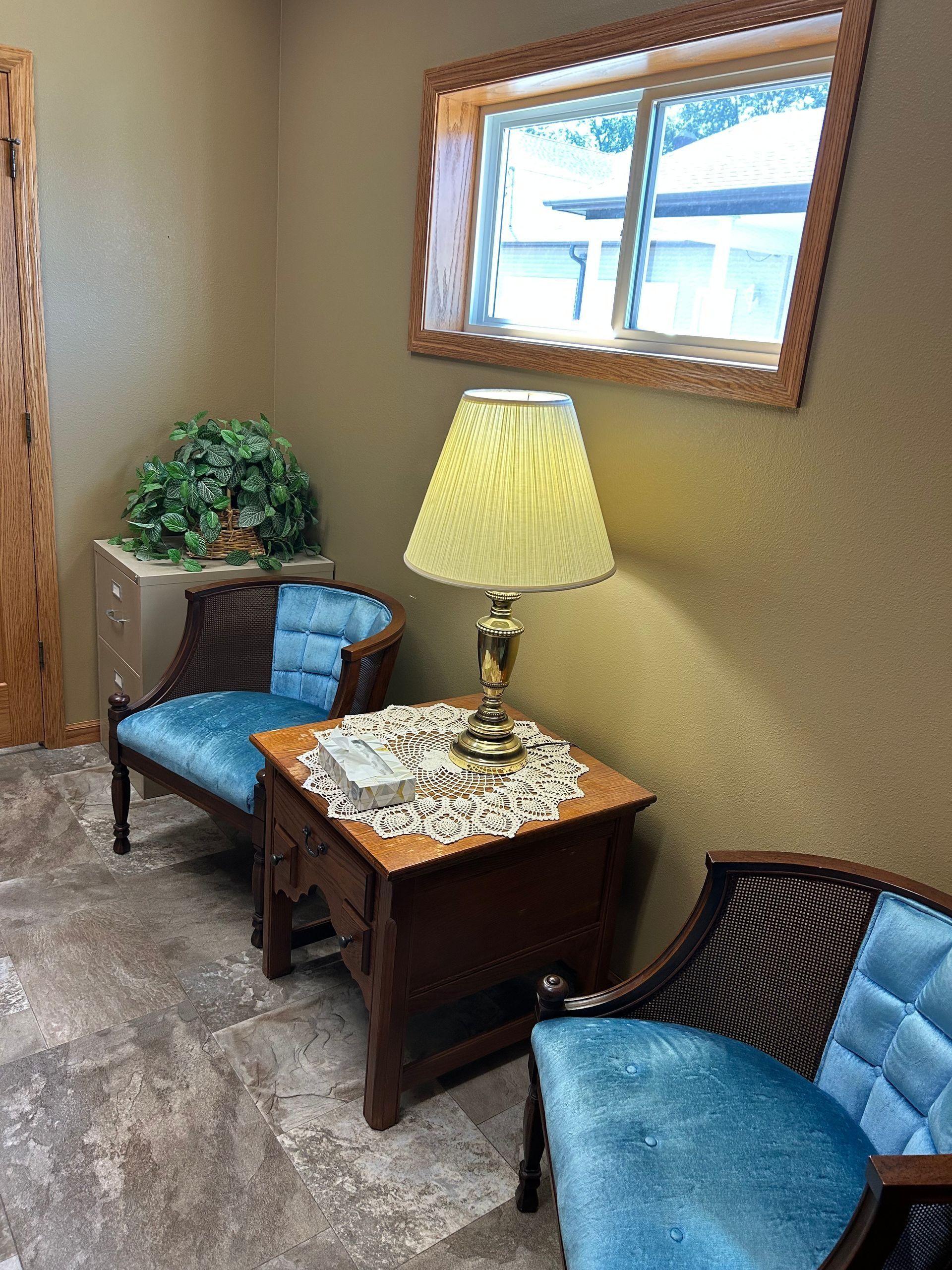Cozy waiting area with two blue chairs, a side table, lamp, plant, and window with wood trim.