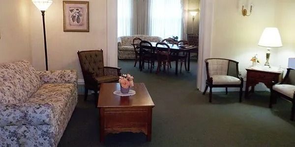 Hotel suite with floral sofa, coffee table, two chairs, lamp, and dining area in the background