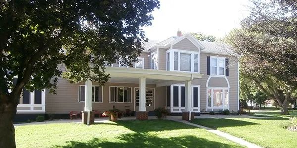 Large two-story beige house with a covered porch and white trim, surrounded by trees and green lawn