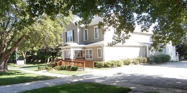 Two-story beige house with white trim, red front steps, and a sunlit driveway under leafy trees