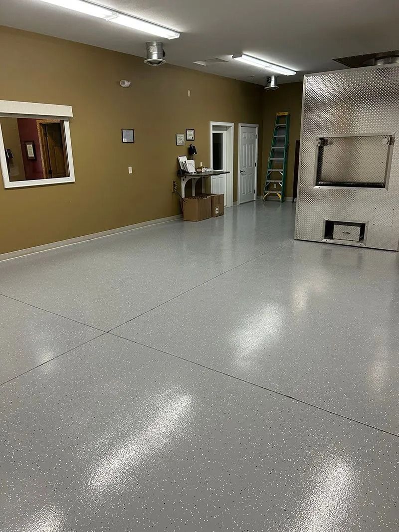 Empty garage-like room with gray speckled floor, tan walls, fluorescent lights, and storage cabinets.
