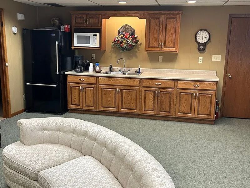 Common room kitchenette with wooden cabinets, black fridge, wall clock, and a beige couch in the foreground