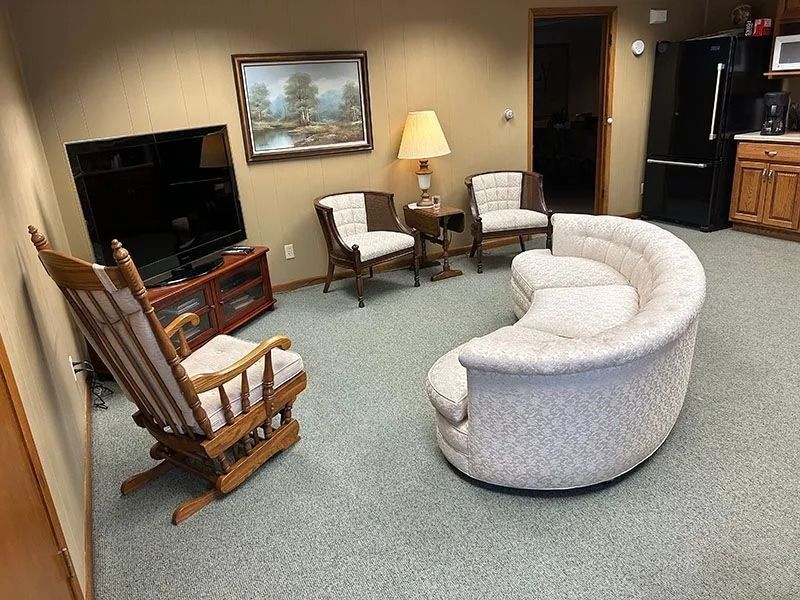 Living room with rocking chair, curved white sofa, TV, lamp, and framed painting on carpeted floor.