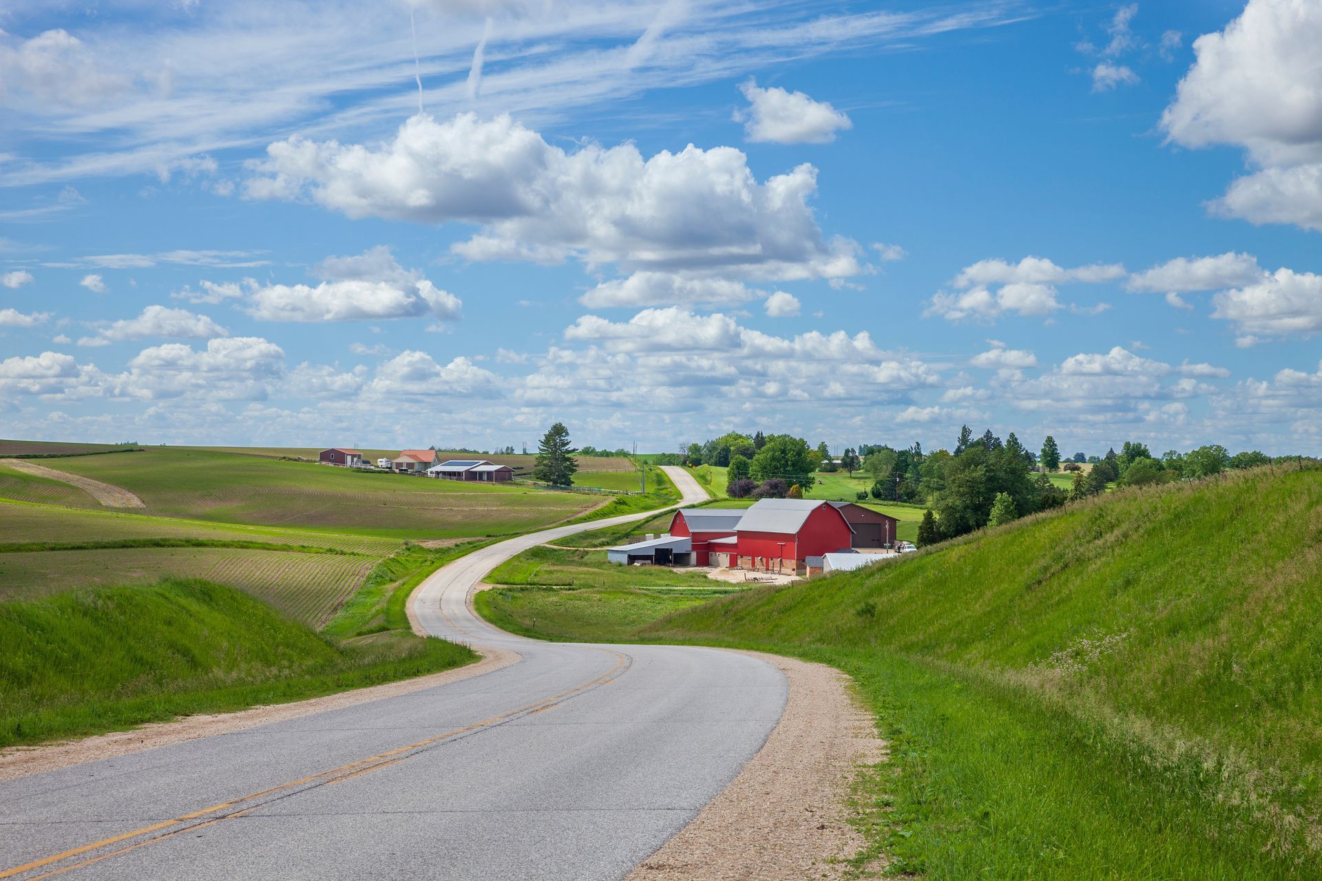 Winding gravel road through green fields to a red farmhouse under a blue sky with clouds