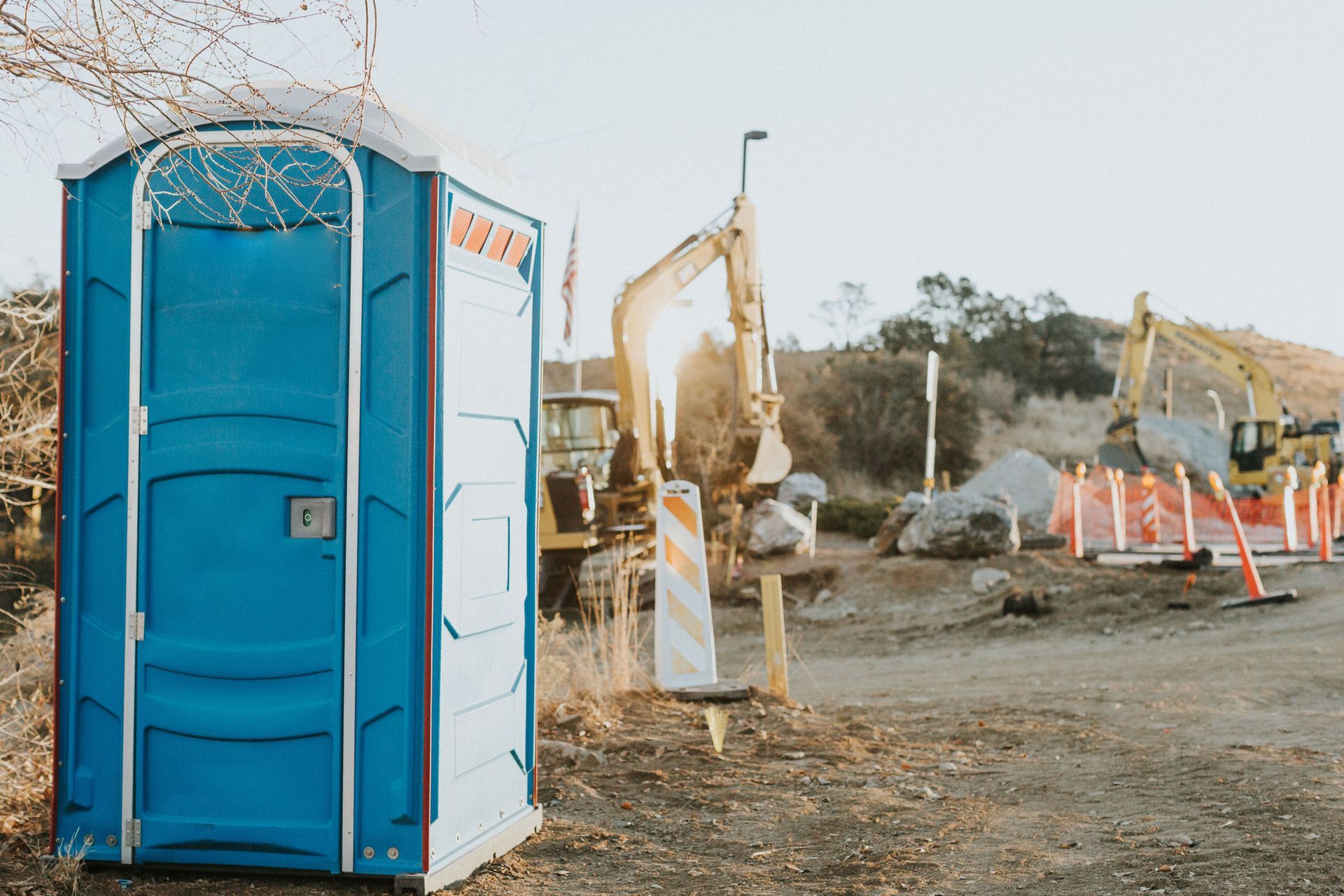 Blue porta-potty sits in a construction site with heavy machinery, safety barriers, and dirt in the late afternoon sun.