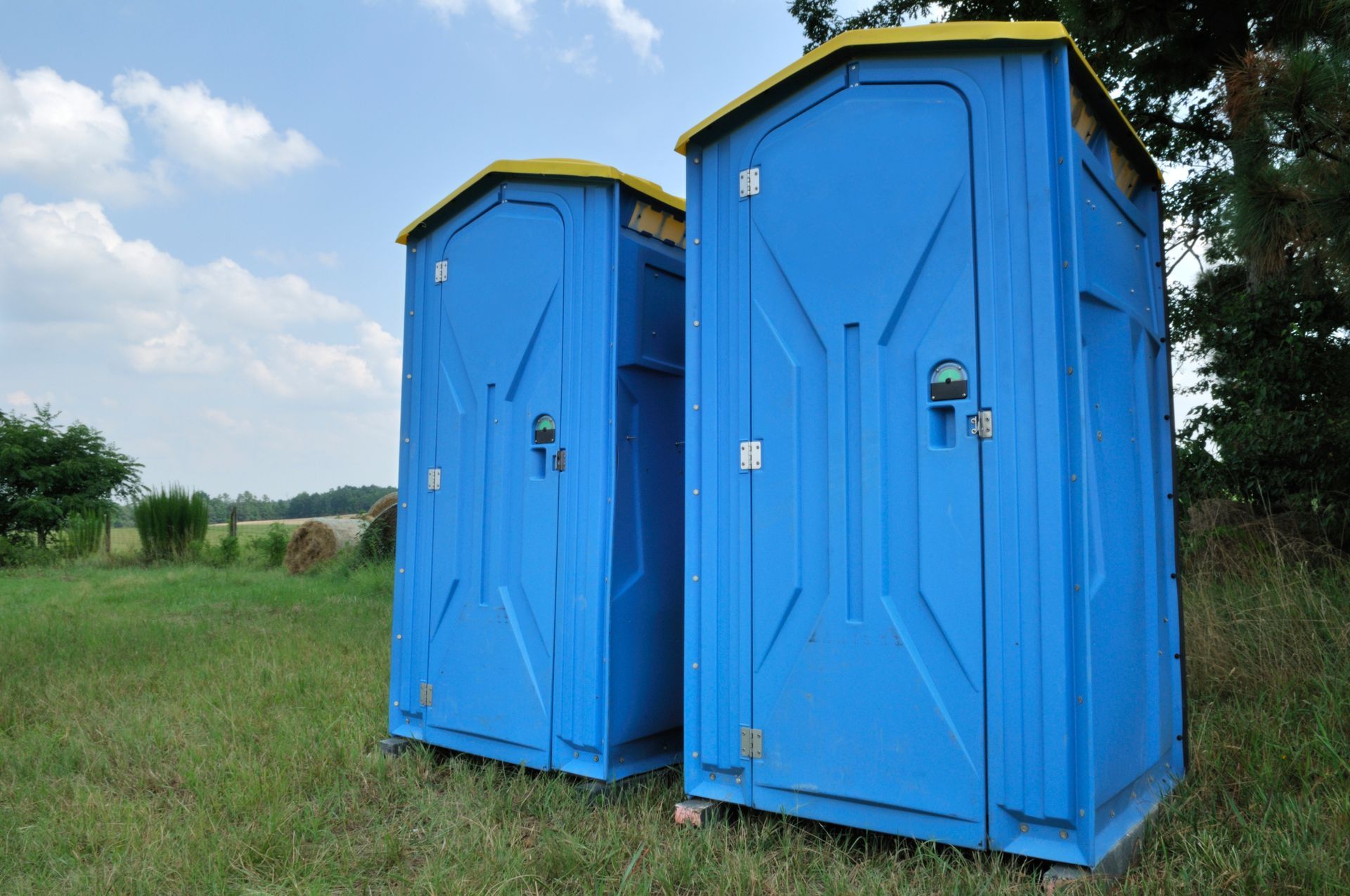 Two blue portable toilets on green grass in a field with a blue sky and trees in the background.