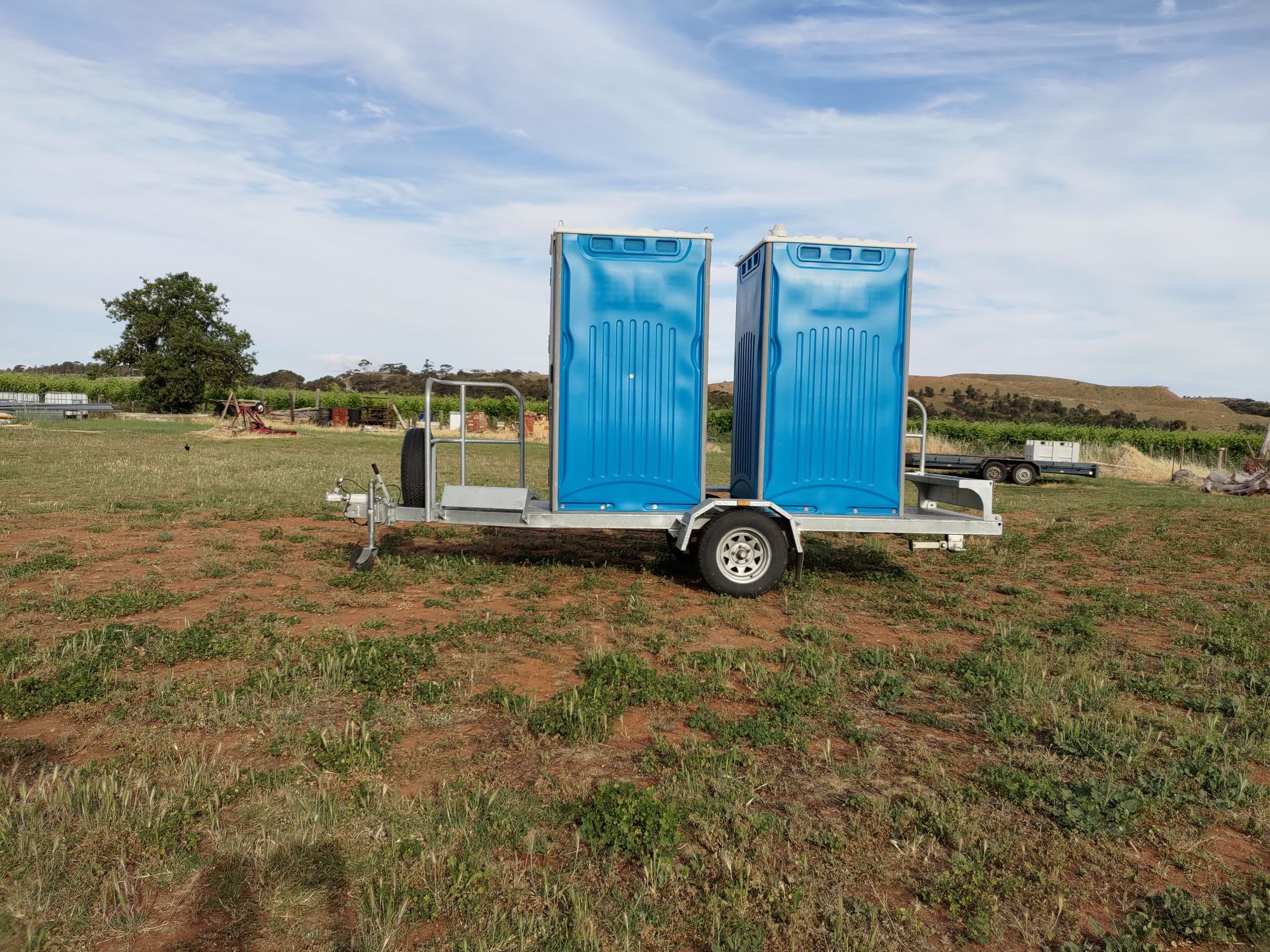 Two blue portable toilets on a trailer in a grassy field under a cloudy sky.