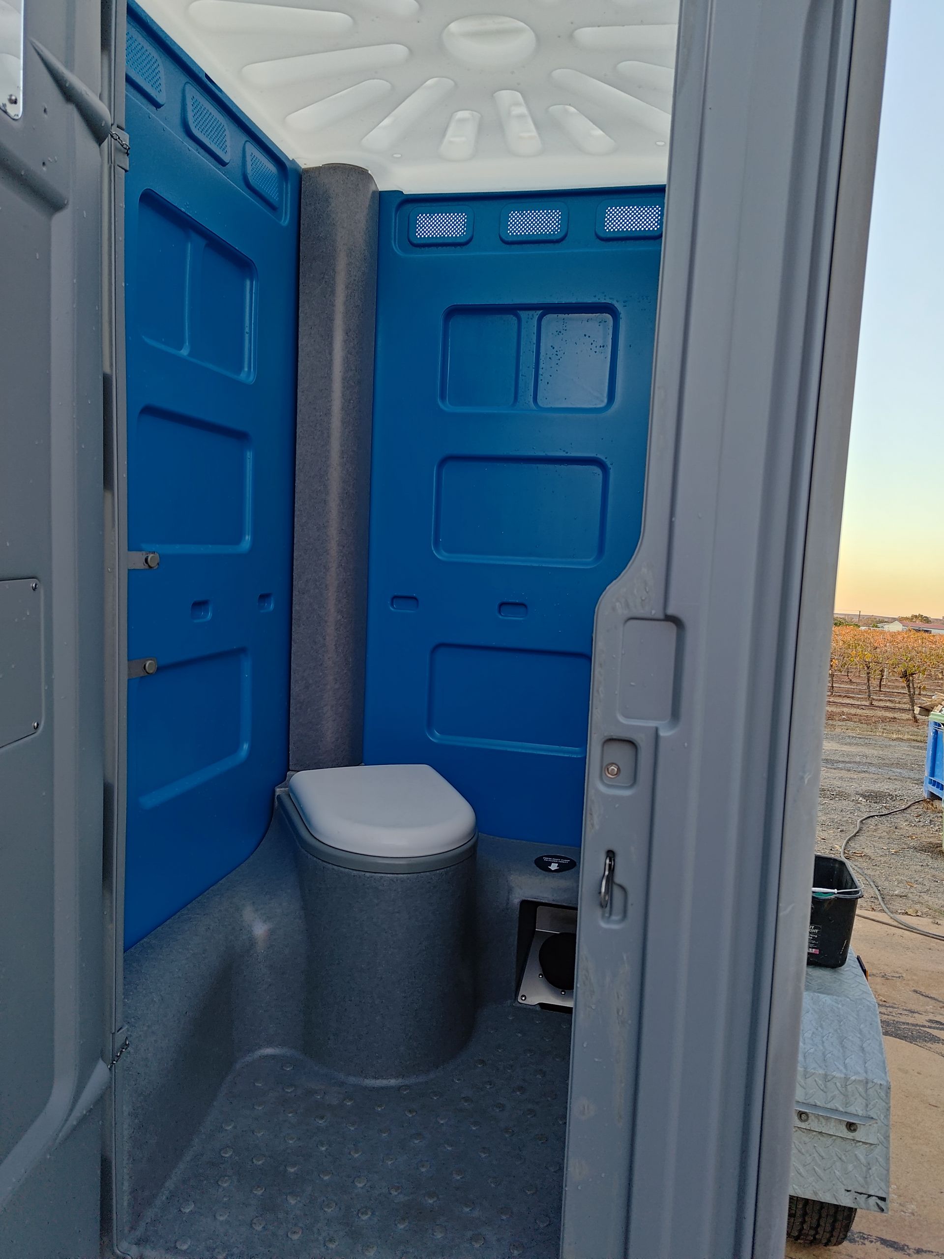 Interior view of a blue and gray portable toilet. Toilet is white. Setting is outdoors.