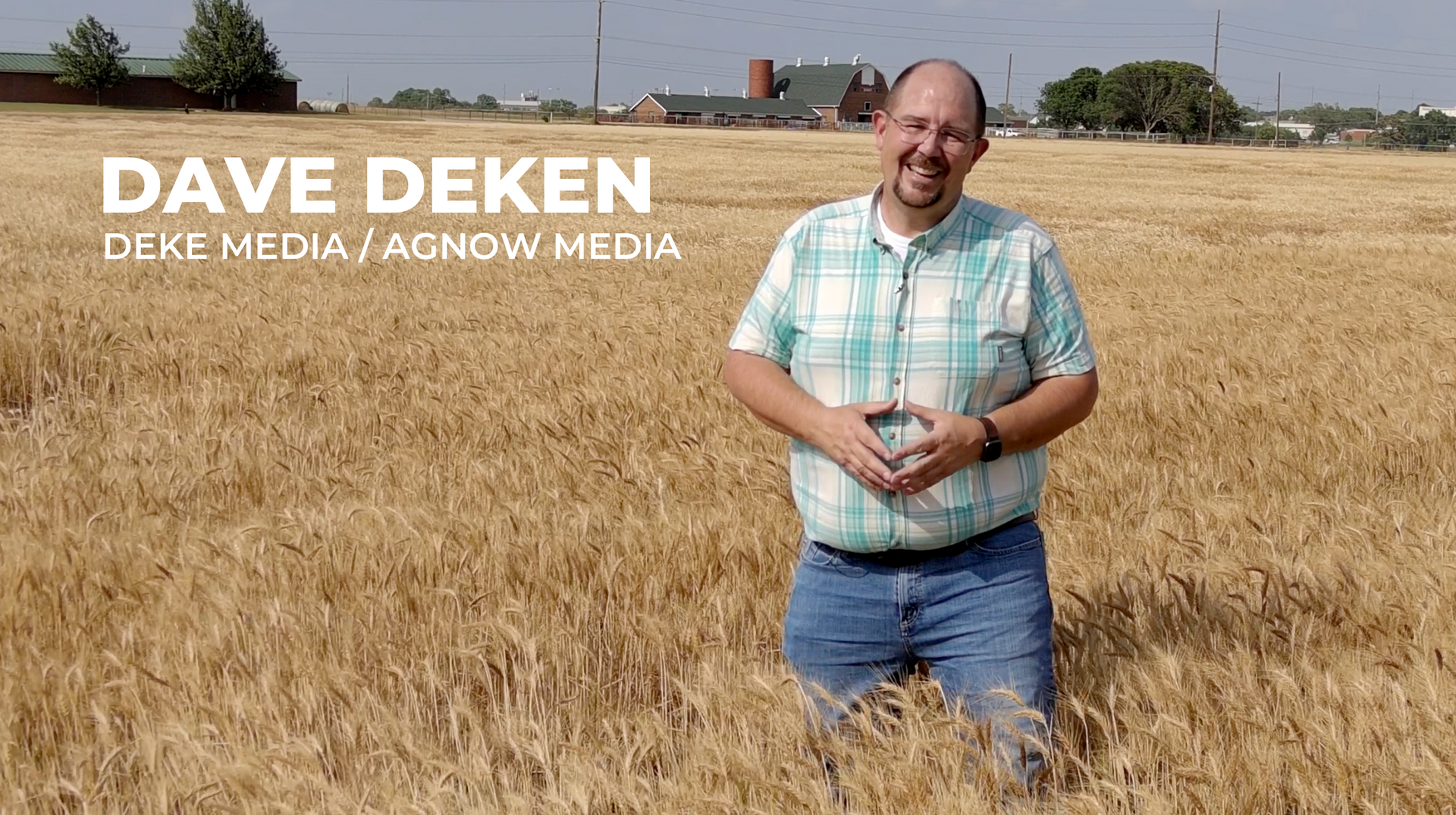 Man standing in a golden wheat field under a clear sky, wearing a plaid shirt and jeans, smiling at the camera
