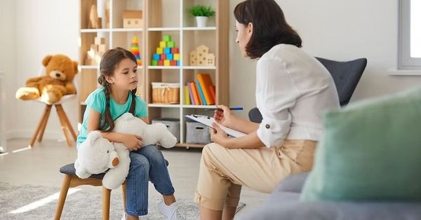 Child and therapist talking in a bright playroom, with toys and stuffed animals nearby.