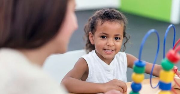 Child smiling at a bead maze across from an adult in a bright classroom