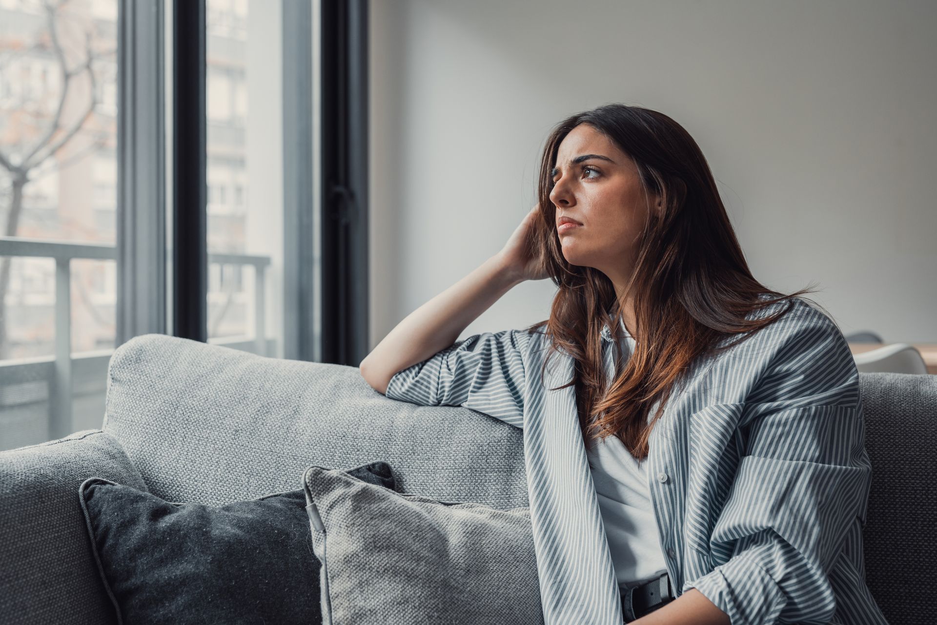 Woman sitting by a window on a gray couch, gazing outside in a softly lit room.