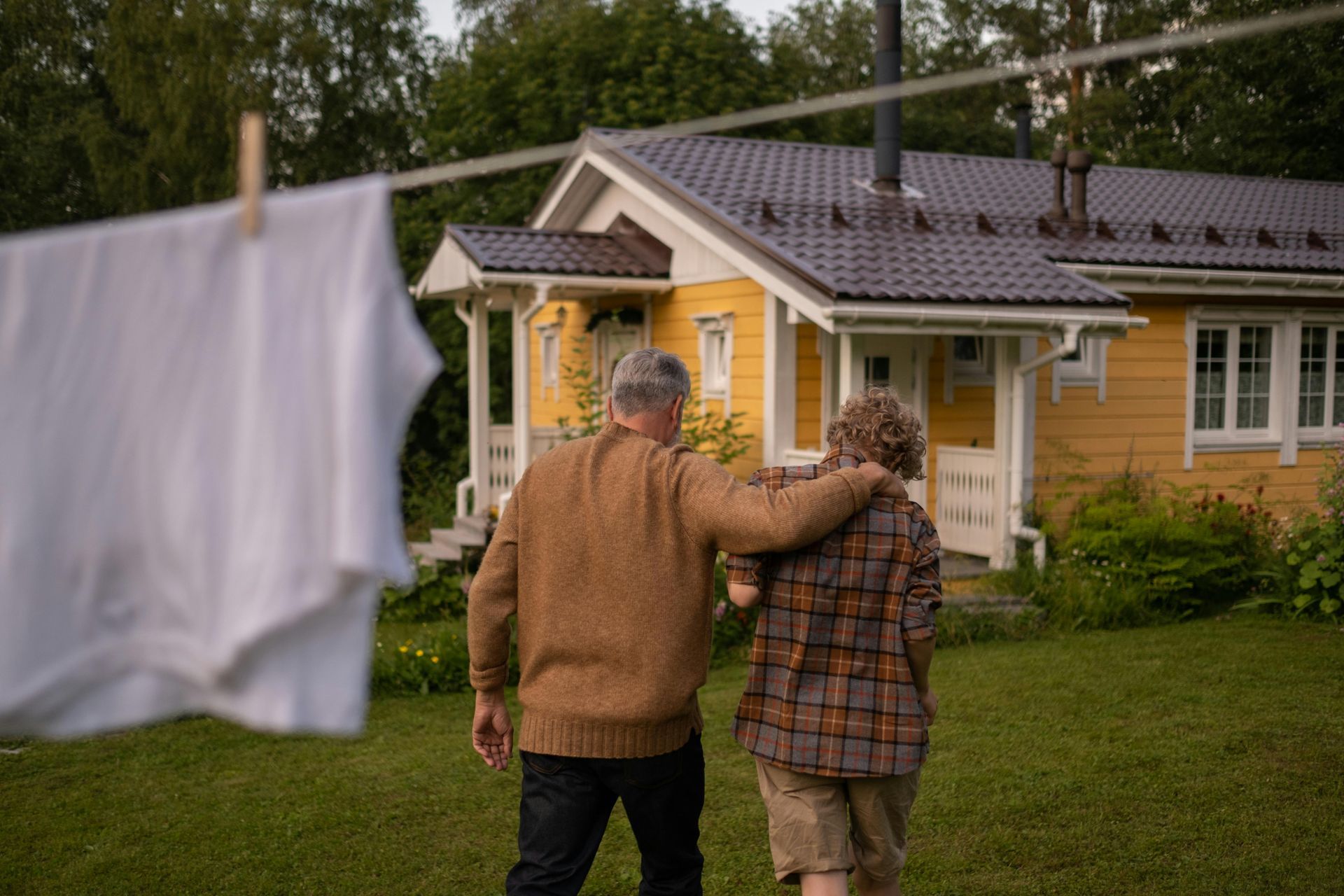 Two people walking arm-in-arm toward a yellow house, with white laundry hanging in the foreground.