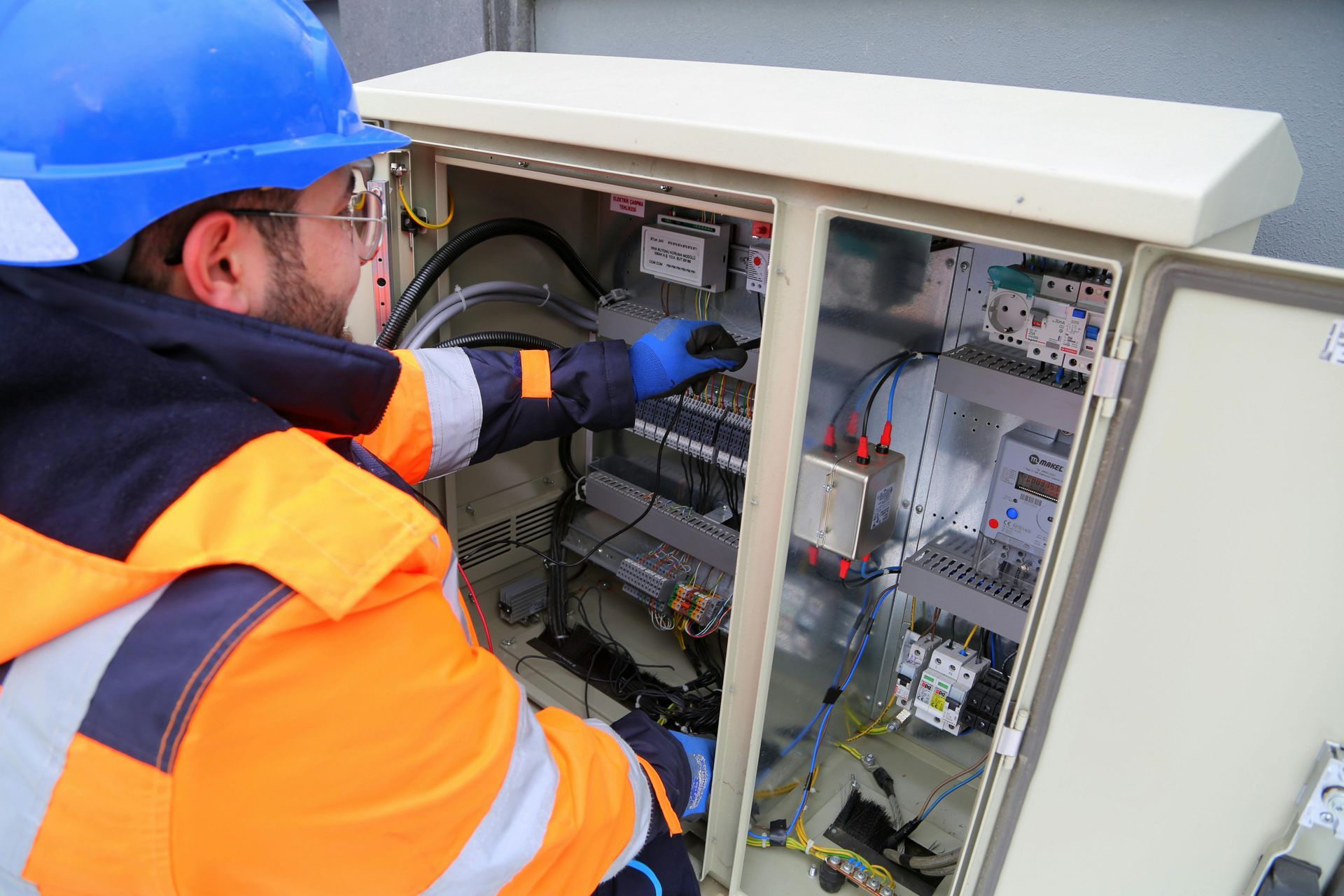 Worker in orange safety gear inspecting wiring inside an open electrical control cabinet