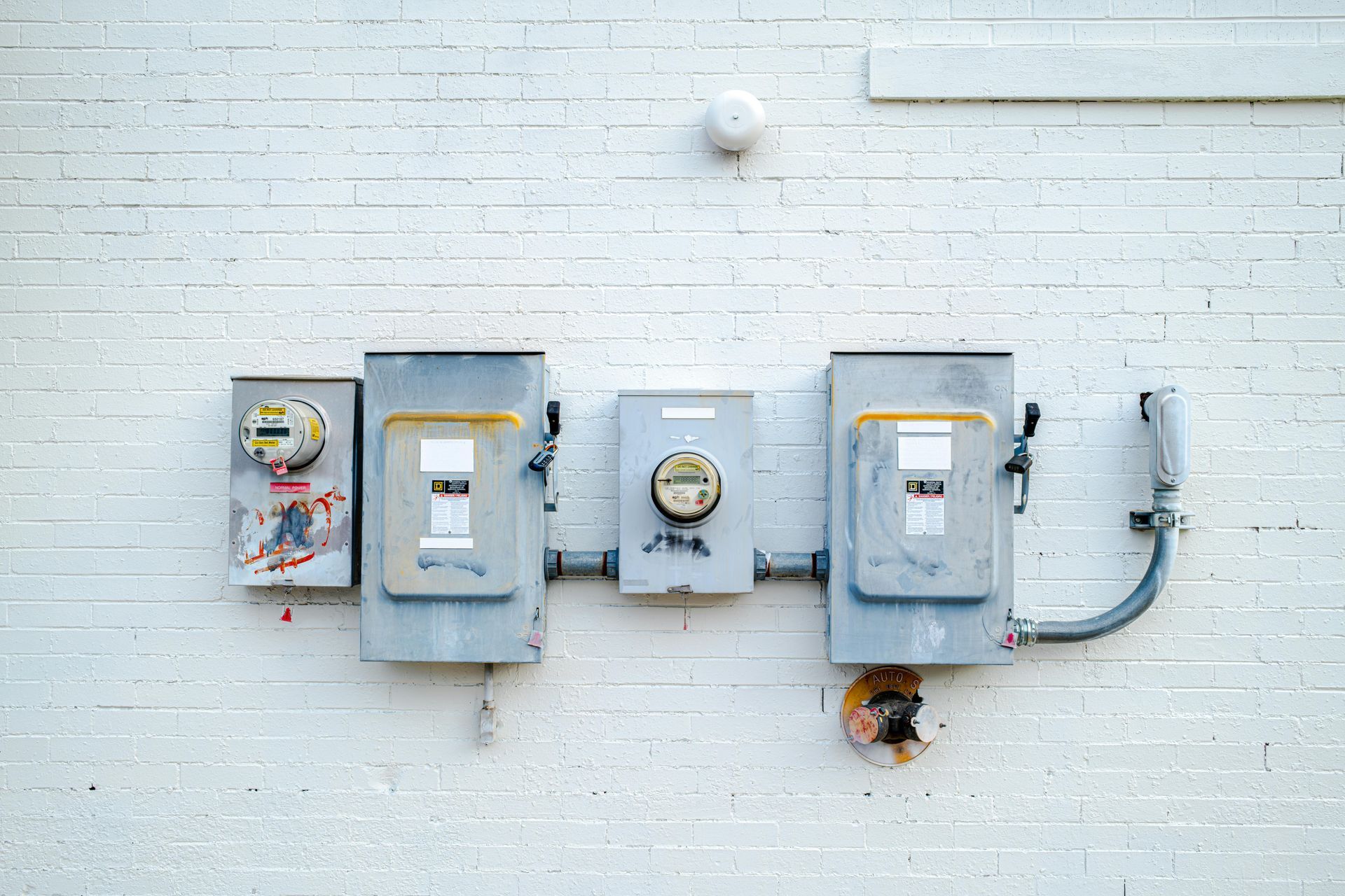 Gray utility meters and electrical boxes mounted on a white brick wall with cables and a small round vent.