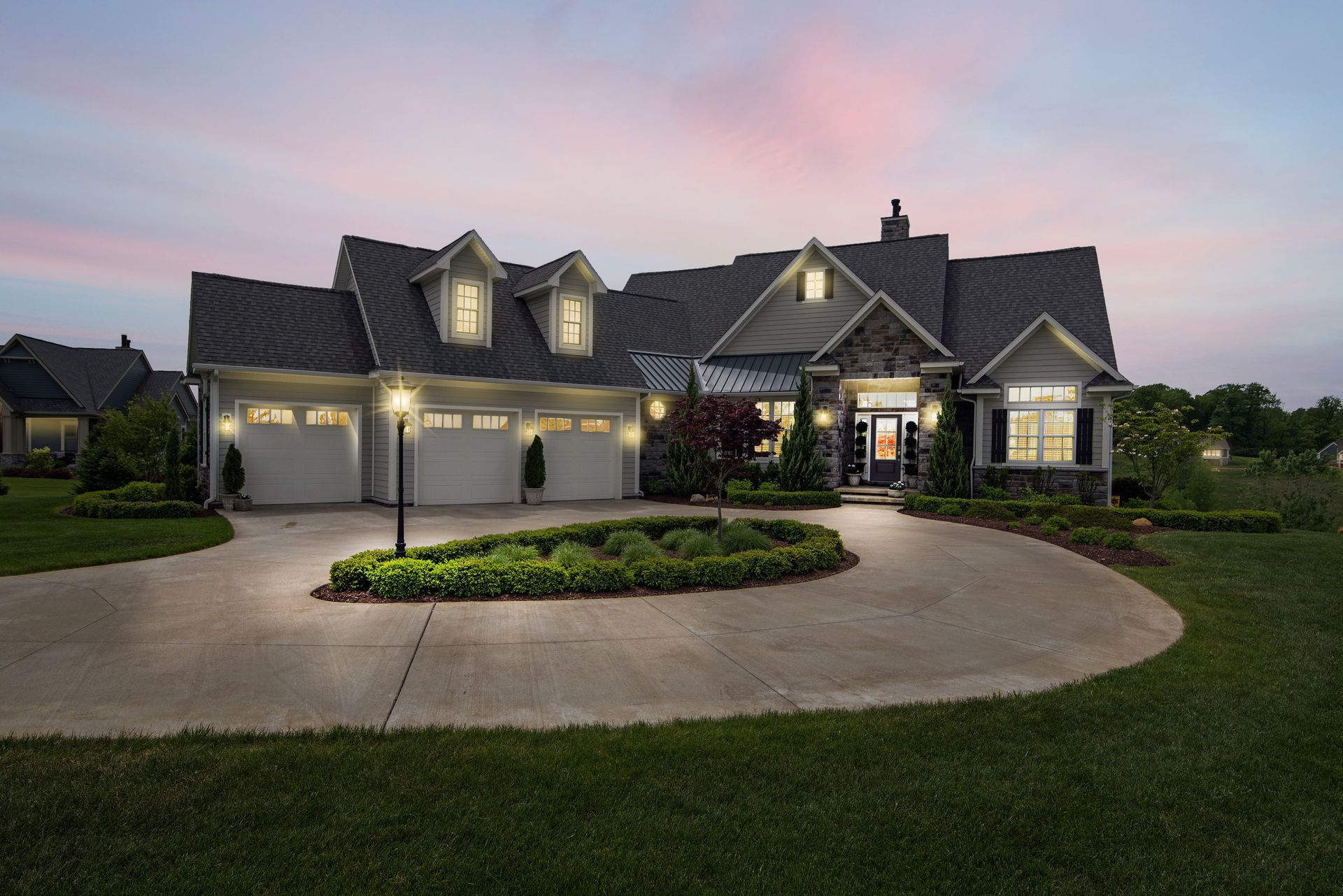 Large suburban house at dusk with lit windows, three-car garage, and a circular driveway around a landscaped island