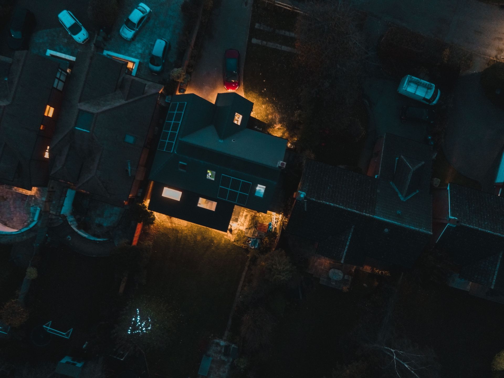 Aerial night view of a lit house surrounded by dark roads and parked cars