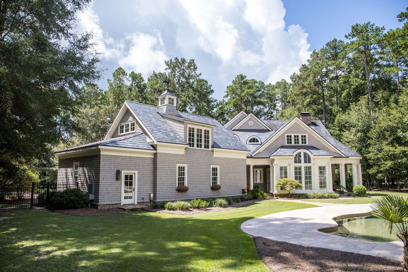 Large gray house with blue roof, surrounded by trees and a pond in a sunny setting.