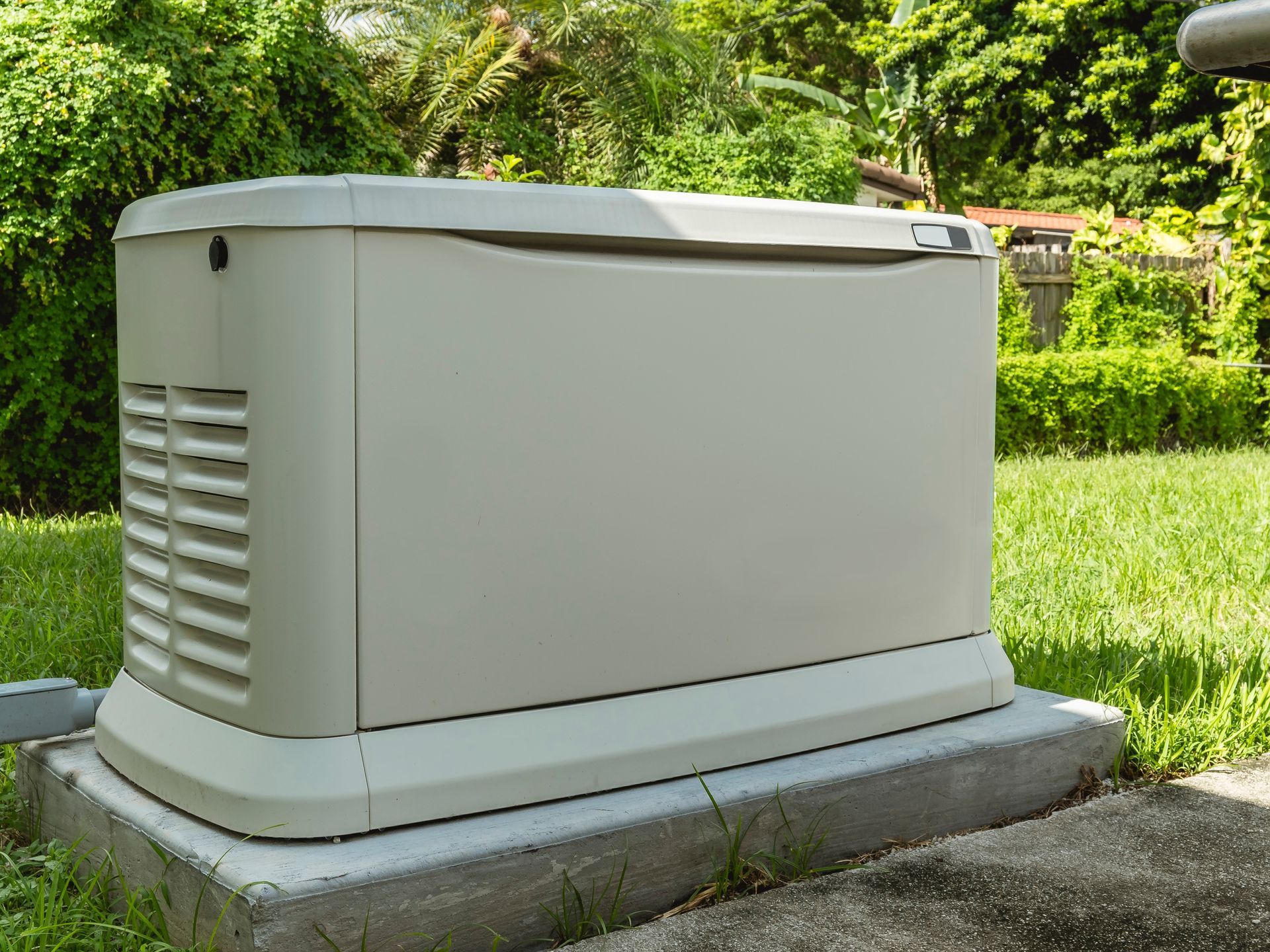Beige home generator on a concrete pad in a grassy yard, surrounded by greenery.