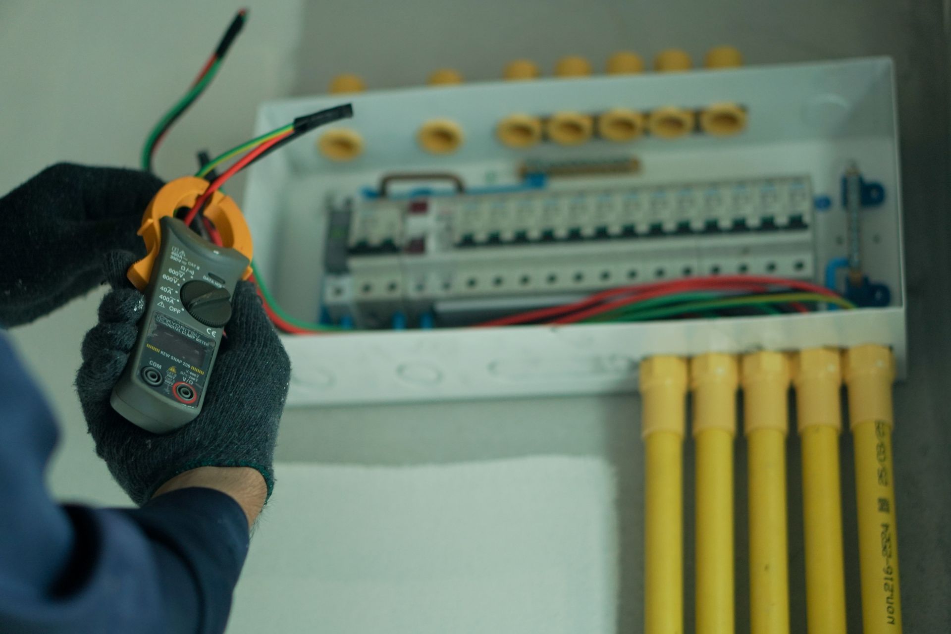 Gloved electrician testing wires in an open electrical panel with yellow conduits nearby