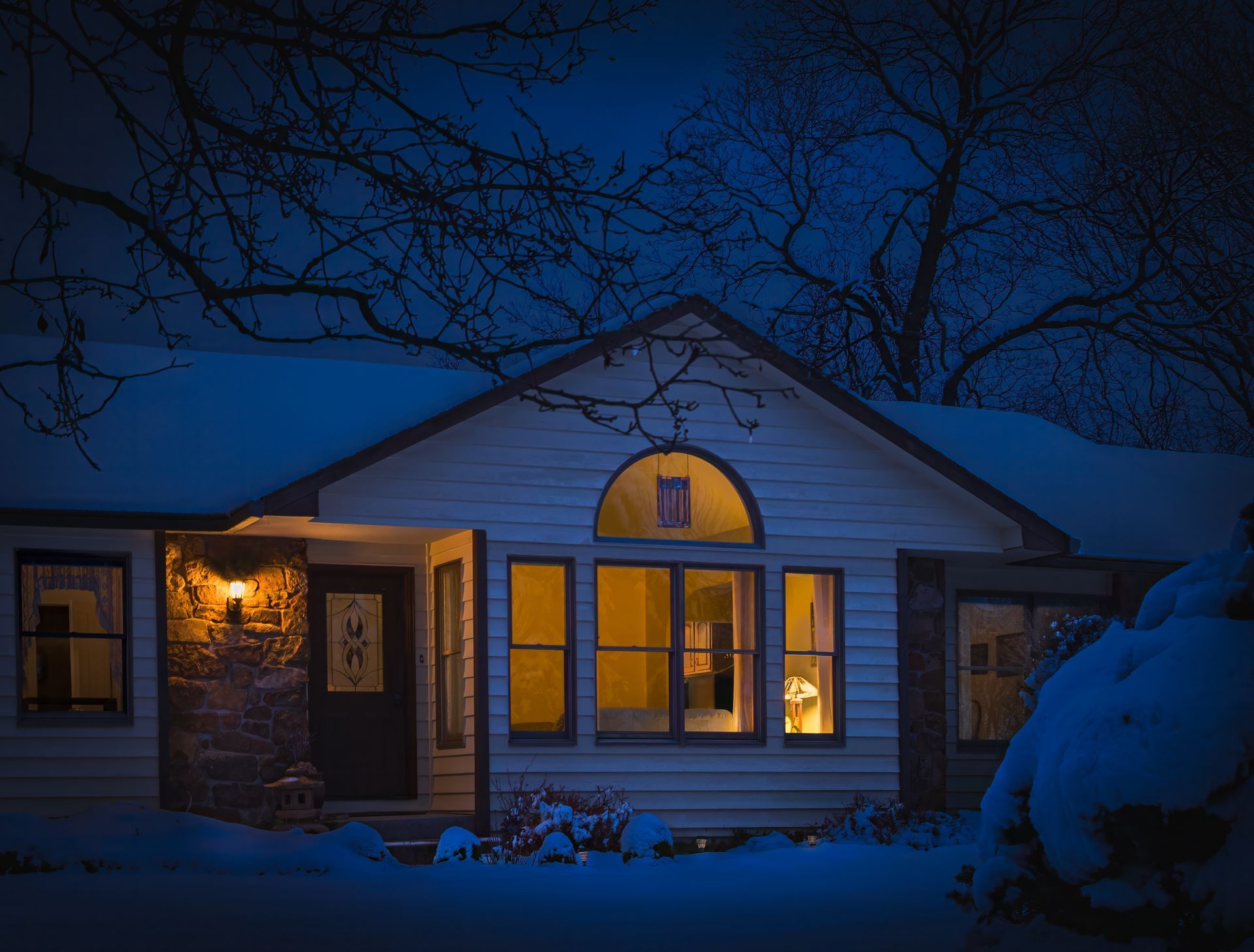Snowy house at night with warm lights glowing in the windows under a dark blue sky