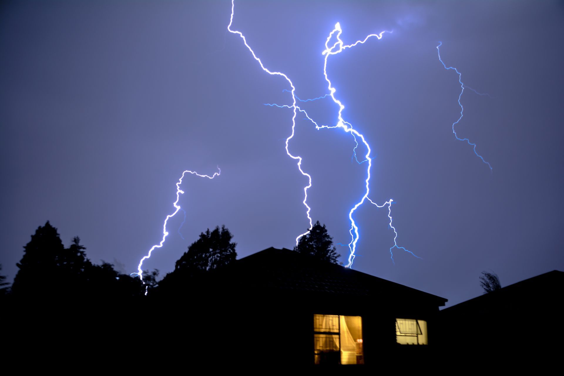 Lightning striking over a dark house at night, with one lit window glowing below.