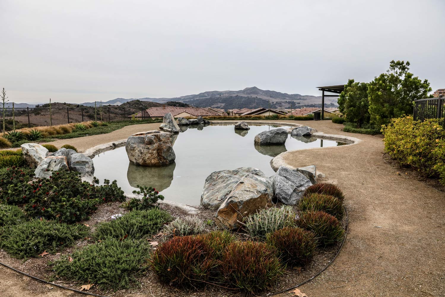 Japanese-style garden with rock pond, gravel paths, shrubs, and distant hills under a cloudy sky