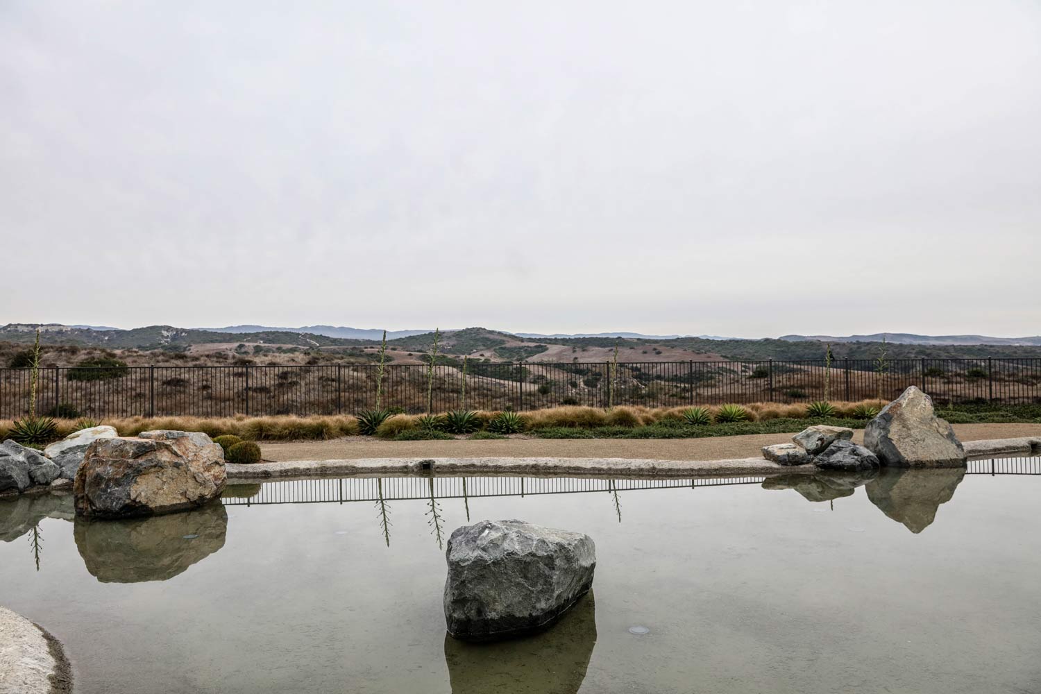 Calm reflecting pool with large rocks, a fence, and a distant cityscape under an overcast sky