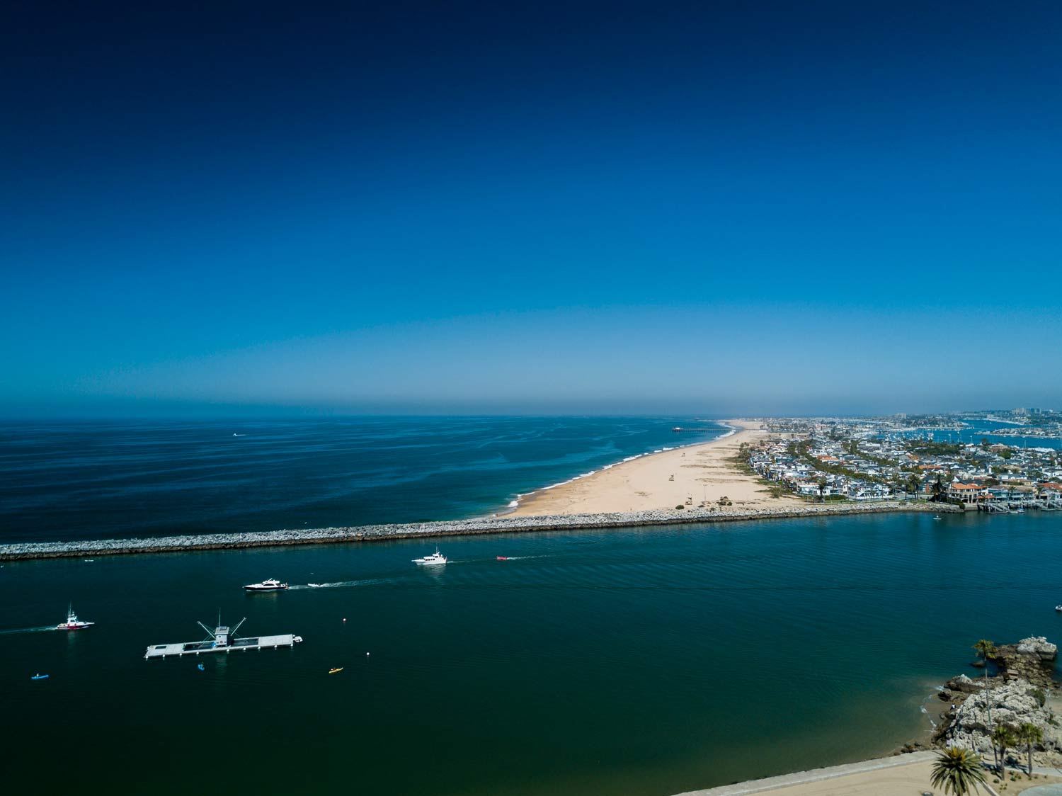 Aerial view of a blue ocean harbor with boats and a sandy peninsula under a clear sky