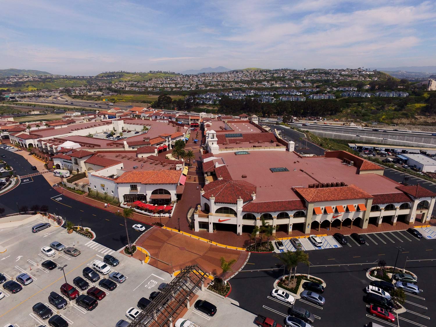 Aerial view of a shopping center with red roofs, parking lots, and surrounding hills under a cloudy sky