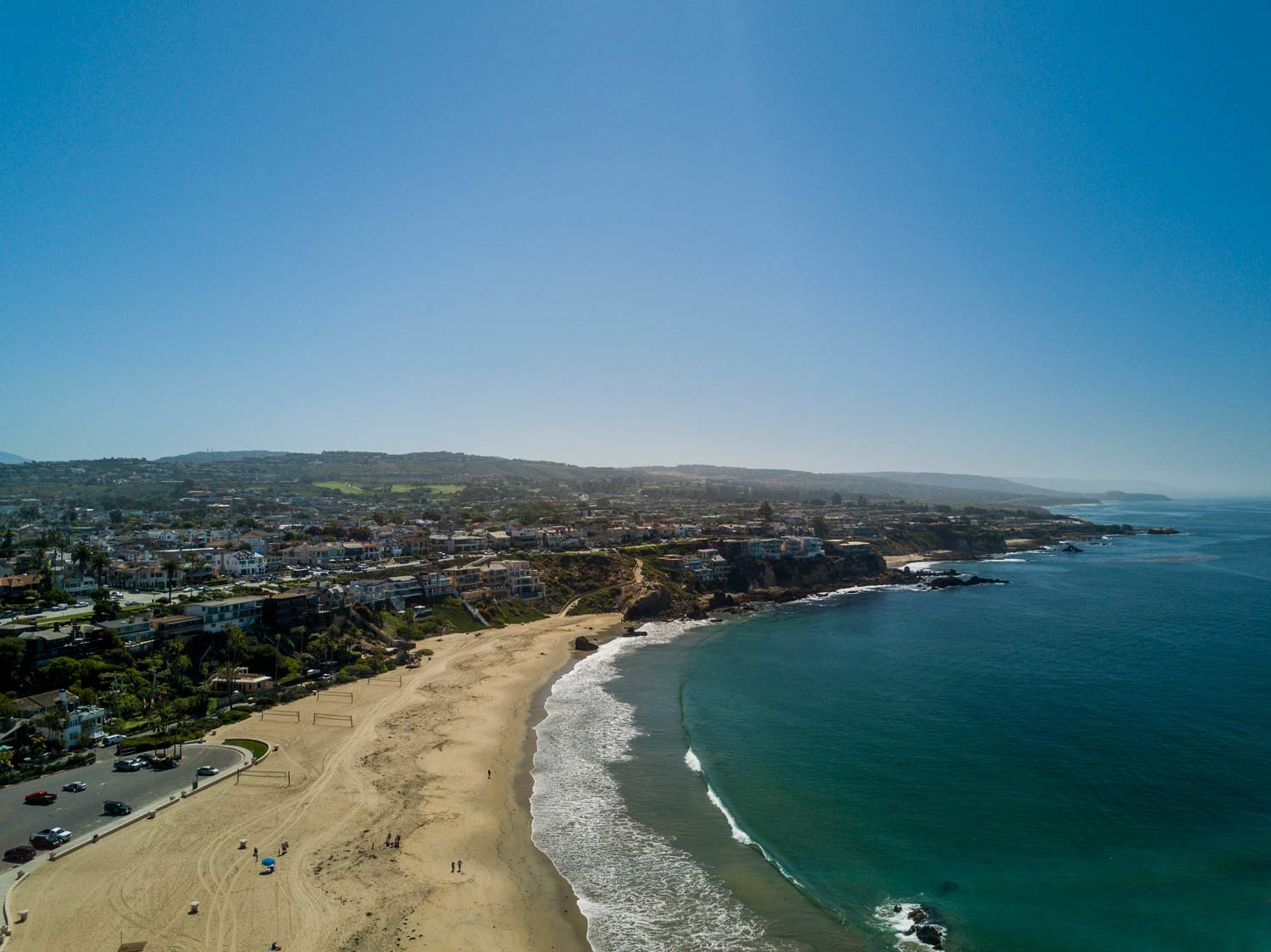 Aerial view of a curving sandy beach beside turquoise ocean and a coastal town under a clear blue sky