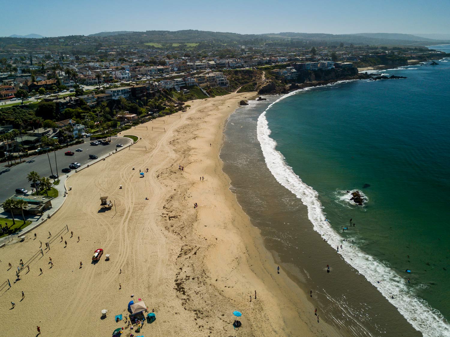 Aerial view of a sandy beach with waves, swimmers, and a coastal city under a clear sky.