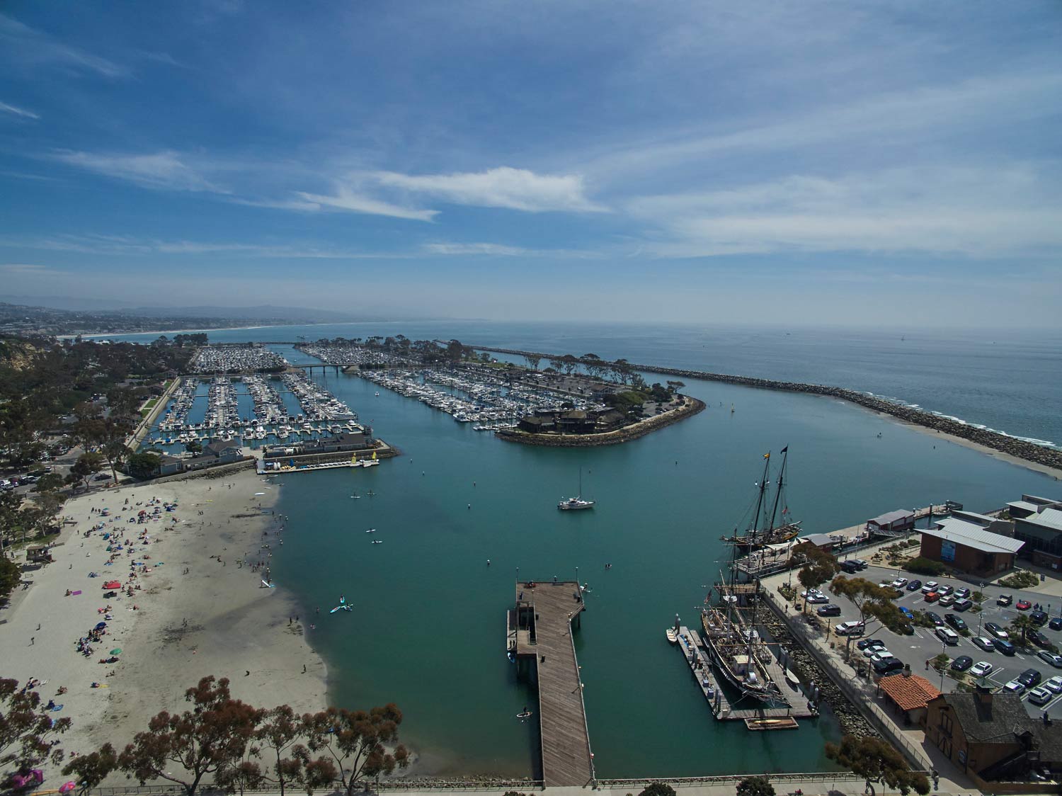 Aerial view of a marina and harbor entrance with boats, sandy beach, and blue water under a clear sky