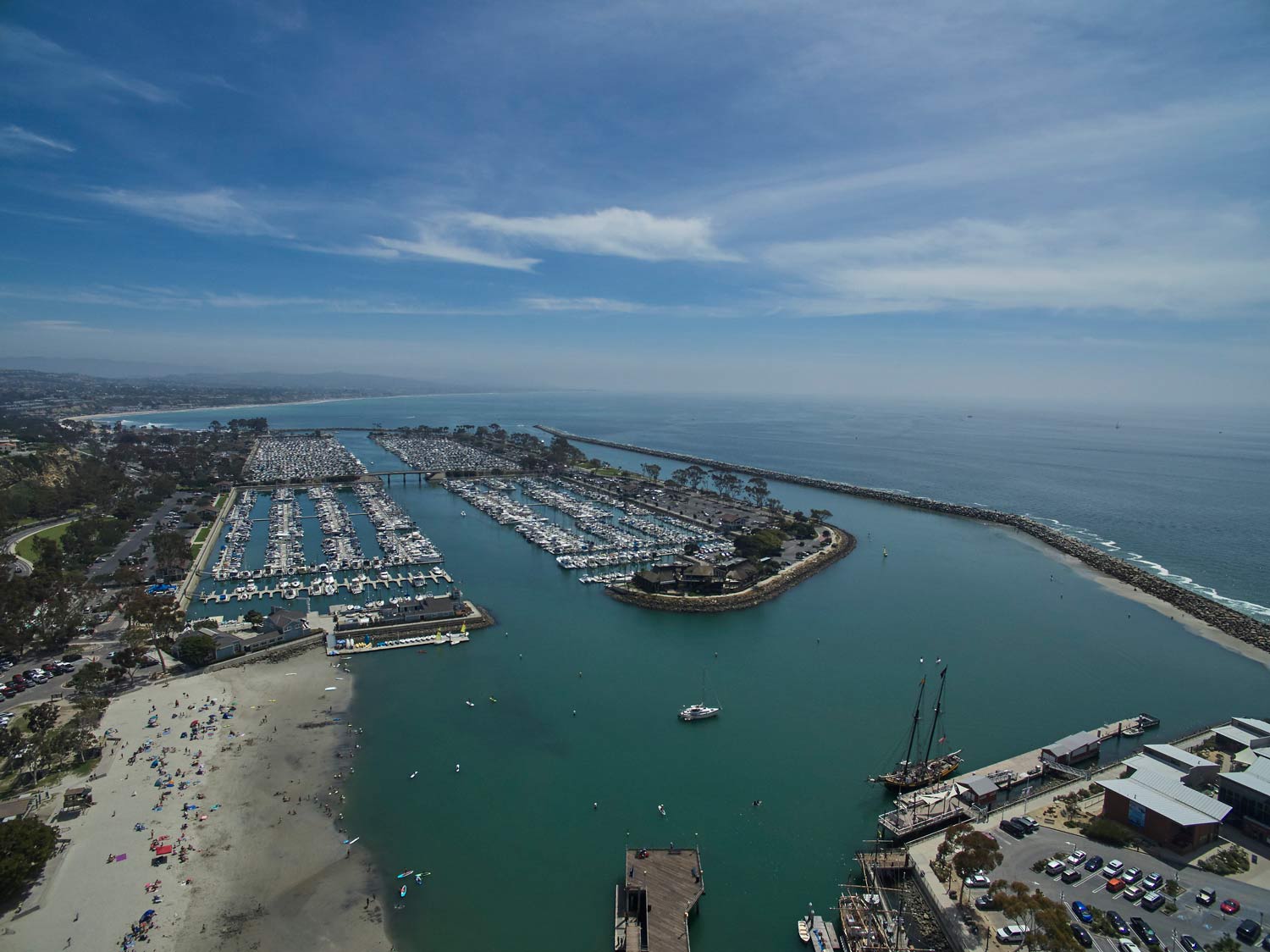 Aerial view of a coastal marina with boats, sandy beach, breakwater, and ocean under a blue sky