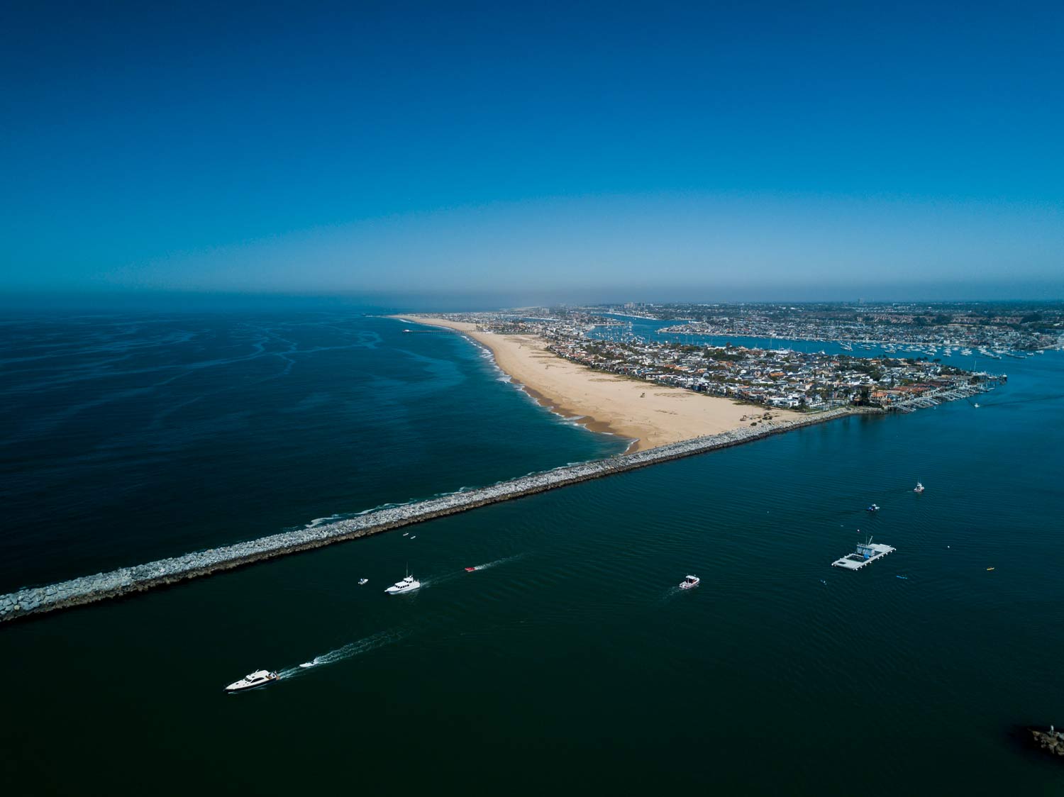 Aerial view of a sandy shoreline between blue ocean and harbor waters with boats and a breakwater