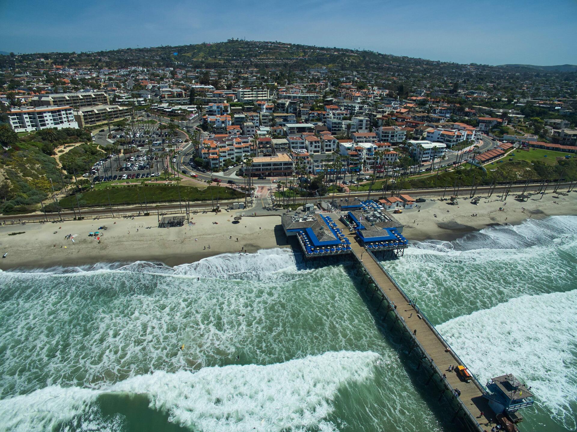 Aerial view of a beach town with a long pier stretching into crashing waves.