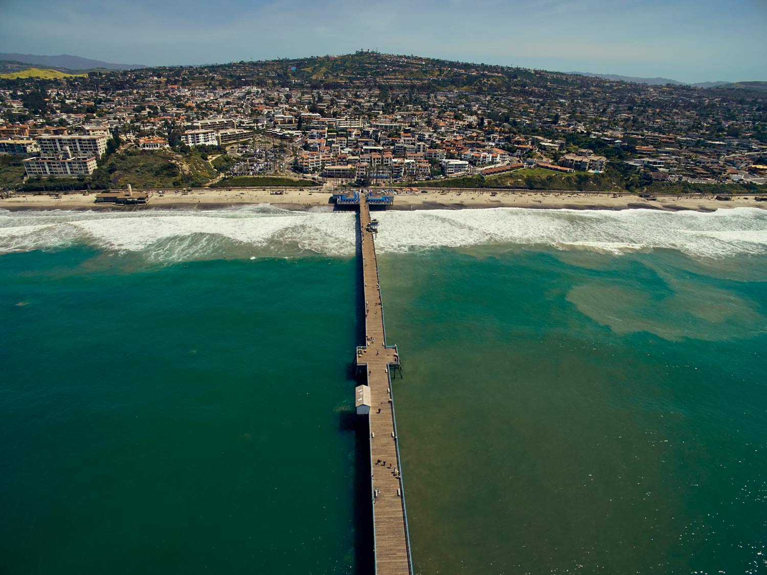 Aerial view of a long pier extending into turquoise water toward a crowded beachfront town and hills