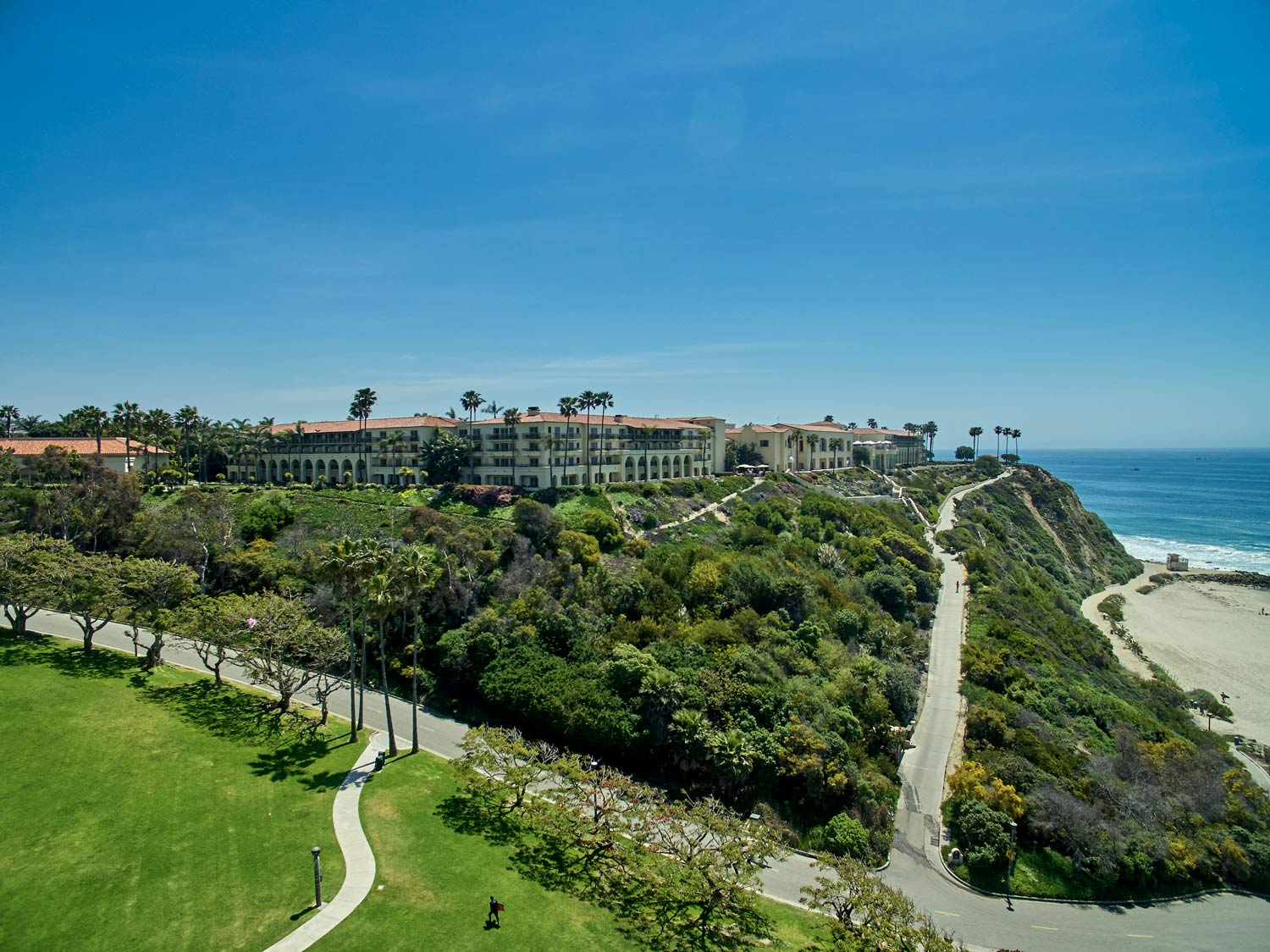 Coastal resort on a bluff above beach and ocean, with green lawns, paths, and a clear blue sky