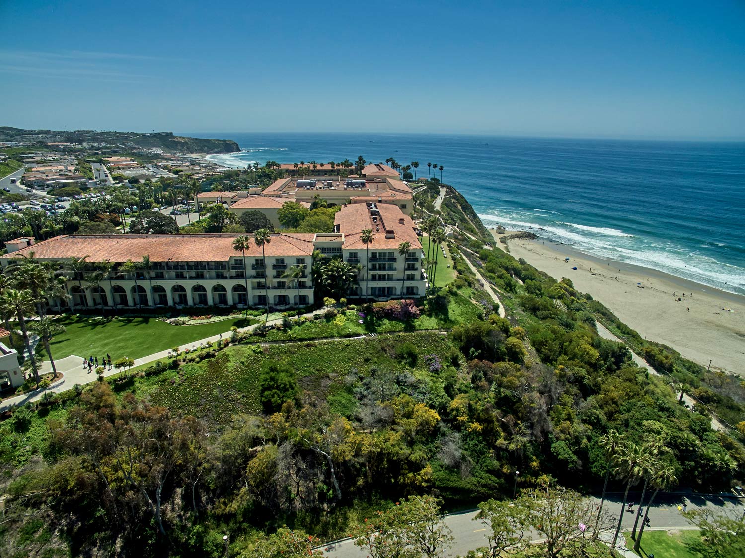 Coastal resort with red-roof buildings beside a sandy beach and blue ocean under clear sky