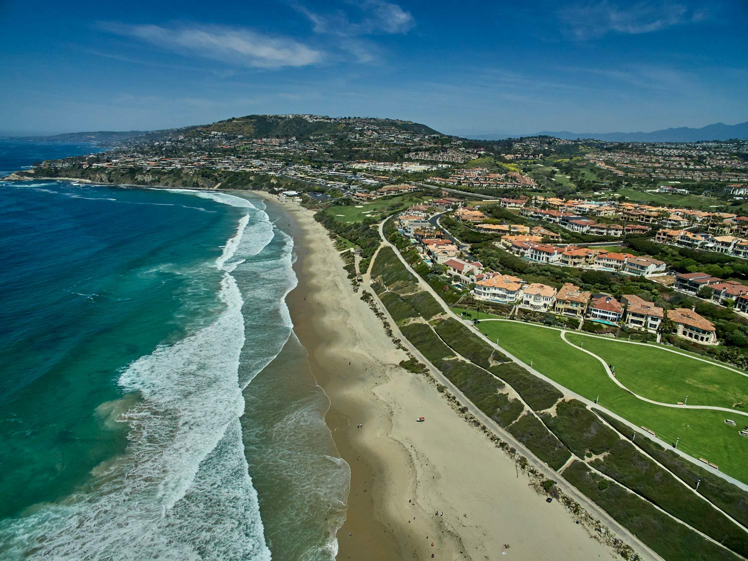 Aerial view of a sandy beach with ocean waves and a coastal town beside green hills.