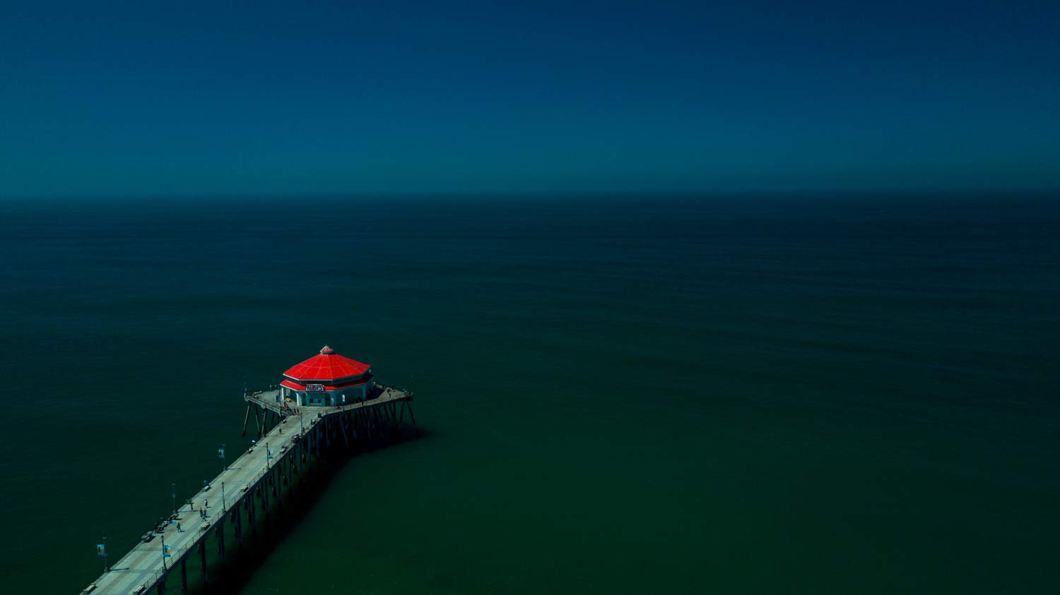 Red-roofed pier extending into a calm, dark ocean under a hazy blue sky.