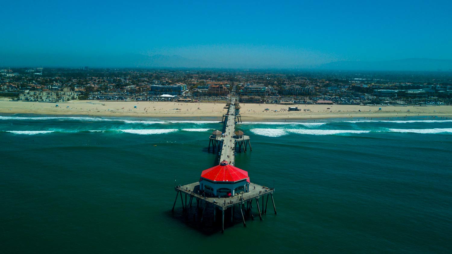 Aerial view of a long pier with a red-roofed pavilion extending into turquoise ocean toward a sandy coastline.