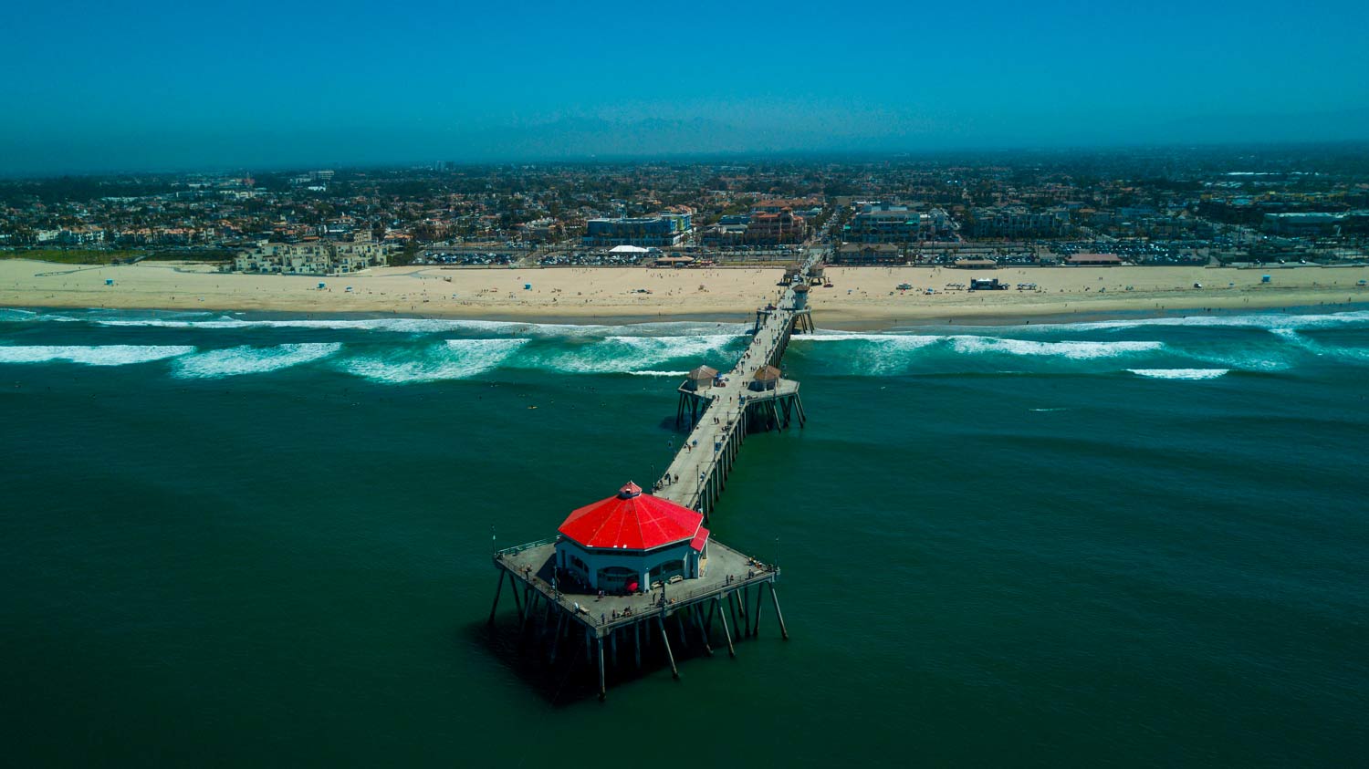 Long pier extending into turquoise ocean toward a sandy beach, with a red-roofed structure at the end