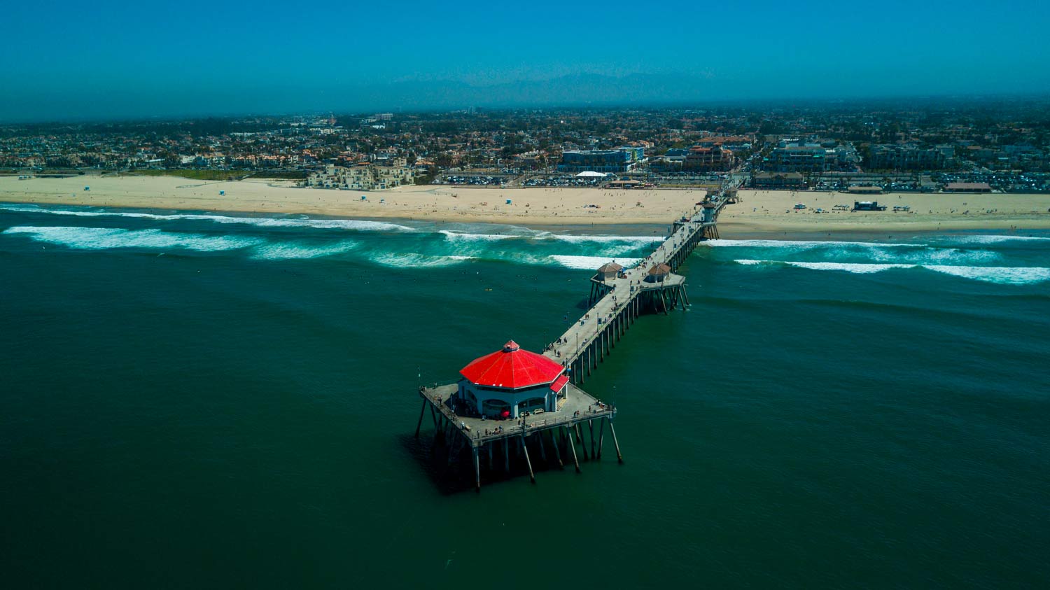 A long pier extends into the ocean to a red-roofed pavilion near a sandy beach and coastline.